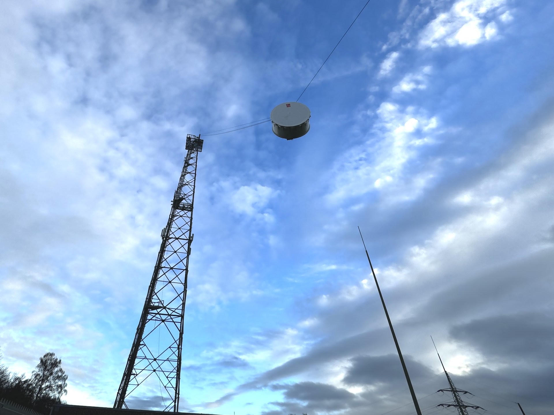 An der Spitze eines 70 Meter hohen Stahlturms ist ein Seil befestigt und schräg bis zum Boden gespannt. An dieser Seilbahn wird eine große Richtfunkschüssel nach oben gezogen. Sie hat die Form einer Trommel mit einem Durchmesser von zwei Metern.